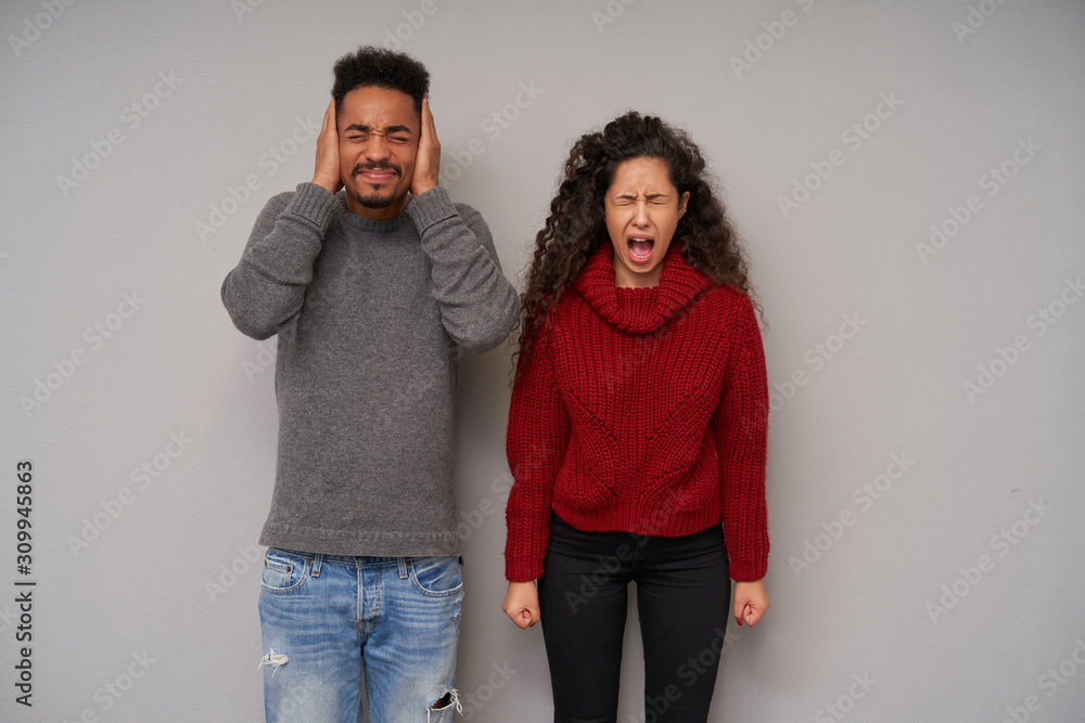 Handsome young dark skinned bearded brunette male frowning face and covering ears with raised palms while his young dark haired curly girlfriend clenching her fists and screaming loud with closed eyes Handsome young dark skinned bearded brunette male frowning face and covering ears with raised palms while his young dark haired curly girlfriend clenching her fists and screaming loud with closed eyes