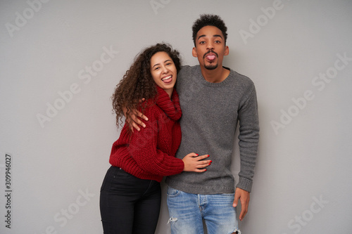 Lovely young cheerful dark skinned couple with curly dark hair hugging each other and fooling while posing over grey background, looking positively at camera and showing tongue