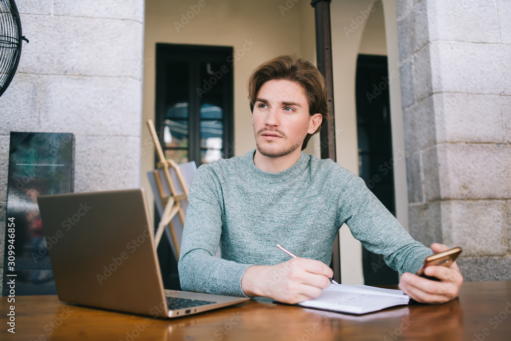 Young freelancer writing in notebook and using smartphone while looking away Young freelancer writing in notebook and using smartphone while looking away