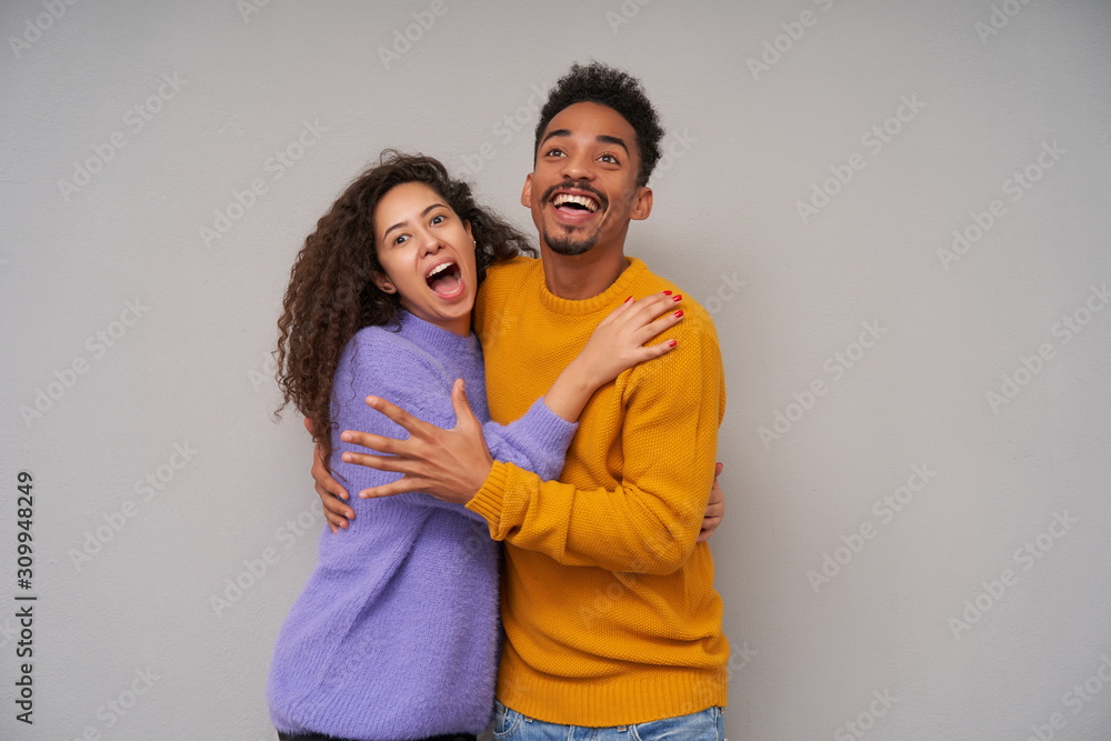 Overjoyed attractive young dark skinned curly couple in casual clothes embracing each other happily and rejoicing about something good happened, isolated over grey background Overjoyed attractive young dark skinned curly couple in casual clothes embracing each other happily and rejoicing about something good happened, isolated over grey background