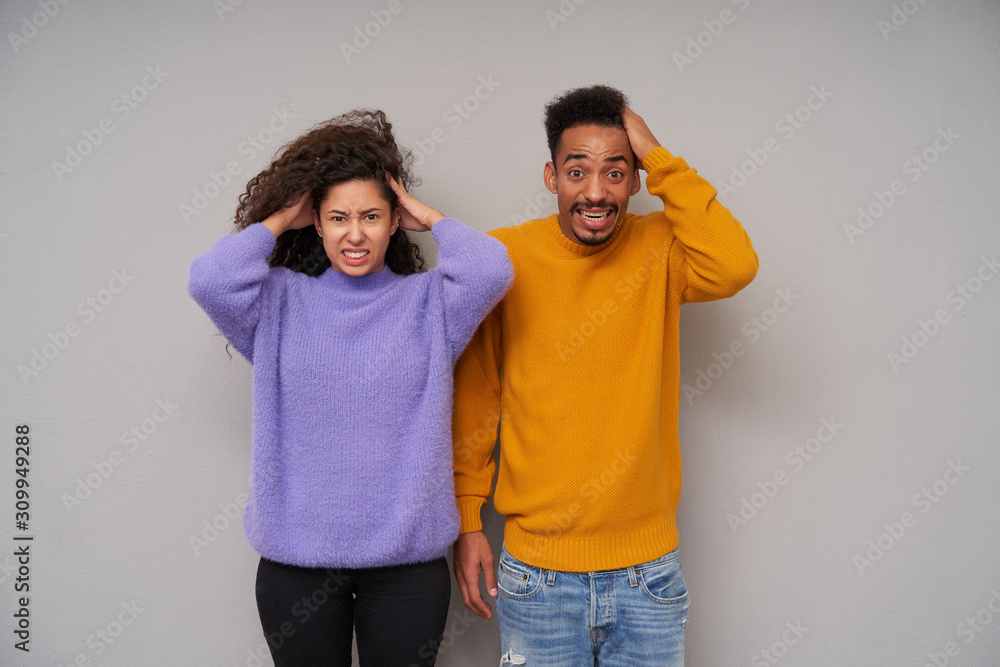 Stressed young dark skinned brunette couple looking to camera with displeased grimace and holding their heads with raised hands, frowning faces while posing over grey background Stressed young dark skinned brunette couple looking to camera with displeased grimace and holding their heads with raised hands, frowning faces while posing over grey background