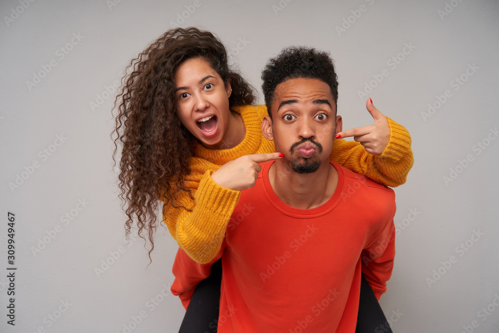 Studio shot of cheerful funny young curly brunette couple fooling and making faces while standing over grey background in casual clothes, spending nice time together on weekend Studio shot of cheerful funny young curly brunette couple fooling and making faces while standing over grey background in casual clothes, spending nice time together on weekend