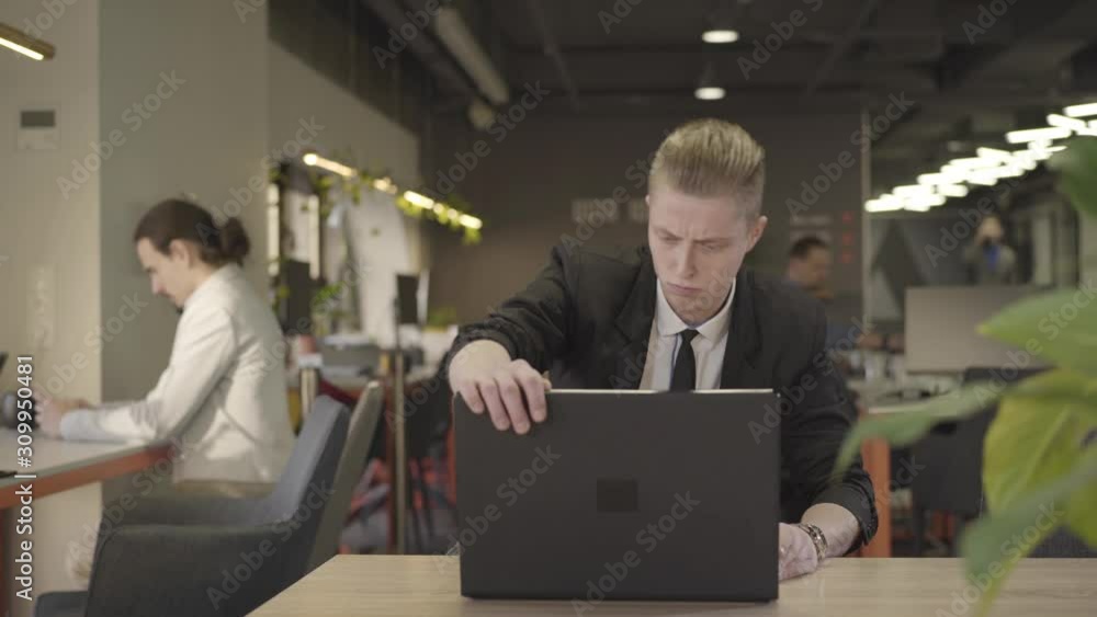 Front view of concentrated redhead Caucasian man sitting on chair at the table and opening laptop. Confident man in formal black suit working in open space office. CEO, management. Front view of concentrated redhead Caucasian man sitting on chair at the table and opening laptop. Confident man in formal black suit working in open space office. CEO, management.