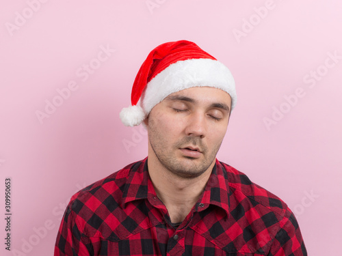 Emotions on the face, tired, holiday hangover, awareness. A man in a plaid rabbit and a Christmas red hat, on a pink background, copy space.