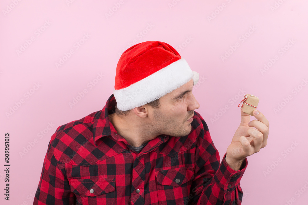 An unhappy, upset man in a red hat, holds a small gift with disgust on his face. Negative human emotions at christmas. Pink background, copy space. An unhappy, upset man in a red hat, holds a small gift with disgust on his face. Negative human emotions at christmas. Pink background, copy space.