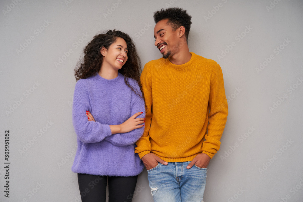 Studio photo of charming young curly long haired lady keeping hands folded on her chest while looking happily on her dark skinned cheerful beared boyfriend, isolated over grey background Studio photo of charming young curly long haired lady keeping hands folded on her chest while looking happily on her dark skinned cheerful beared boyfriend, isolated over grey background