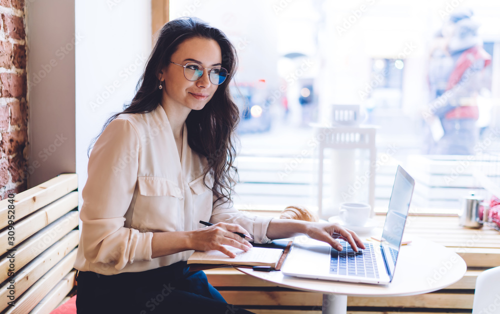 Pleased elegant lady looking away while sitting at table with notepad and laptop in cafe Pleased elegant lady looking away while sitting at table with notepad and laptop in cafe