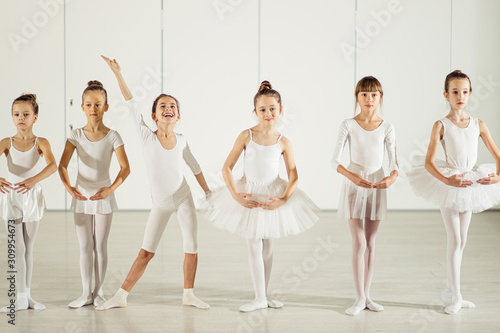 sweet caucasian little girls ballerinas stand in a row in a position wearing white tutu suit, while one girl standing in funny pose, smile, happy ballet group of young girls