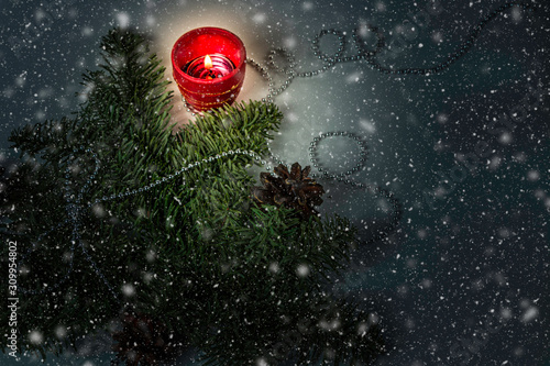 Christmas background with spruce branch, cone, lighted red candle and small beads. Falling snow in foreground. Darkened fabulous atmosphere.