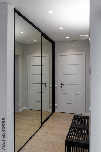 Part of the corridor of a residential house. .There is a door at the end of the room, on the right - a black bench, on the left - wardrobe with mirror that can be seen reflecting the view of the room.