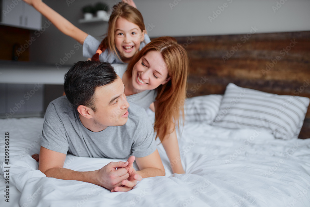 portrait of beautiful happy family lying on bed together, mother father and kid girl kissing hugging and laughing. indoors portrait of beautiful happy family lying on bed together, mother father and kid girl kissing hugging and laughing. indoors