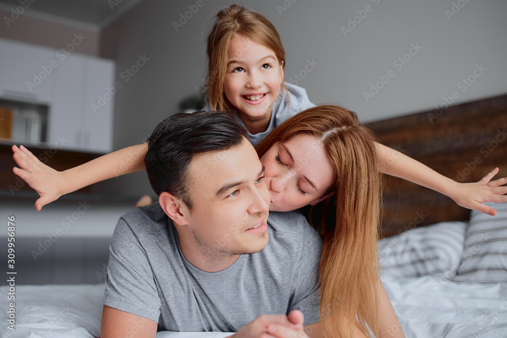 portrait of beautiful happy family lying on bed together, mother father and kid girl kissing hugging and laughing. indoors portrait of beautiful happy family lying on bed together, mother father and kid girl kissing hugging and laughing. indoors