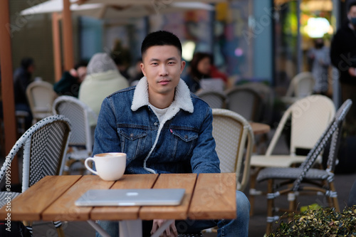 One handsome Asian young man sitting in sidewalk cafe looking at camera. Blur background