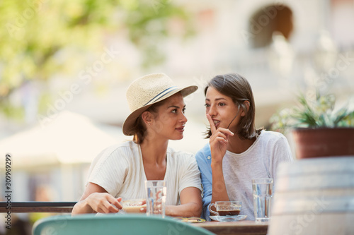  Two beautiful caucasian girls sitting in cafe outside and telling secrets. On table are cups of coffee and grasses with water. Summer time.