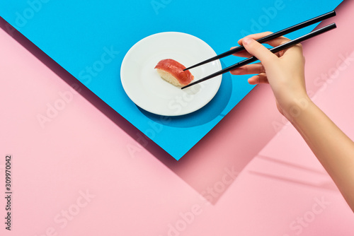 Cropped view of woman eating fresh nigiri with tuna with chopsticks on blue, pink background