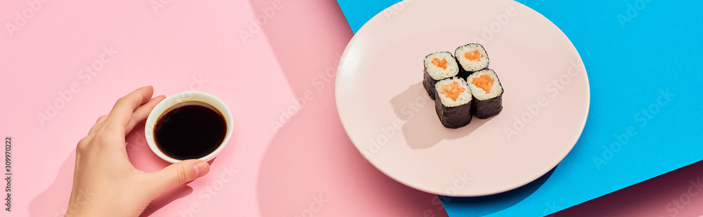 cropped view of woman holding soy sauce near fresh maki with salmon on blue, pink background, panoramic shot
