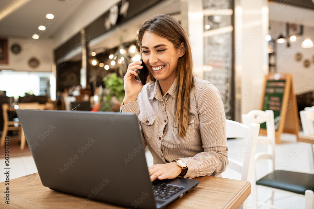 Cheerful attractive young caucasian brunette smiling and talking on her smart phone, She is also browsing on her laptop while sitting in a coffee shop. Cheerful attractive young caucasian brunette smiling and talking on her smart phone, She is also browsing on her laptop while sitting in a coffee shop.