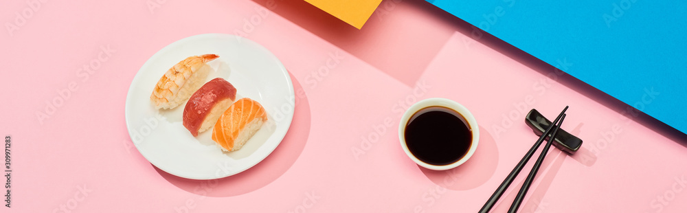 fresh nigiri with salmon, tuna and shrimp near soy sauce and chopsticks on blue, pink, orange surface, panoramic shot