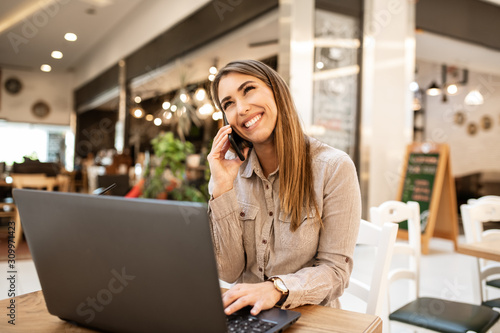 Cheerful young businesswoman with brown hair taking a break from work on her laptop. She is smiling and talking with friend on smart phone. She is sitting inside of a cafe.