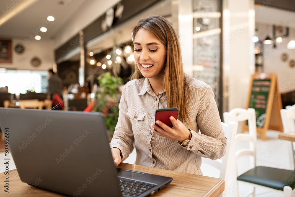Pretty young caucasian woman just finished a conversation and holding her smart-phone. She is typing on her laptop and chatting with her friends. She's sitting in a cafe. Pretty young caucasian woman just finished a conversation and holding her smart-phone. She is typing on her laptop and chatting with her friends. She's sitting in a cafe.