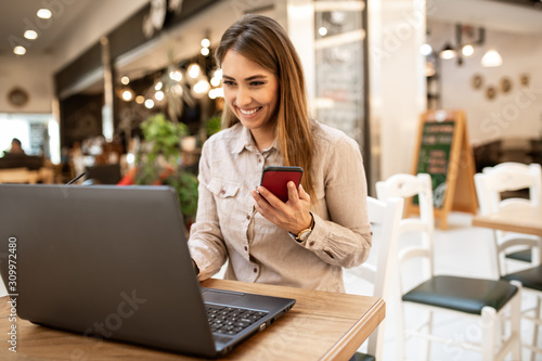Beautiful young caucasian woman with brown hair smiling while browsing on her laptop and checking emails. She is holding her smart phone and sitting in a cafe.