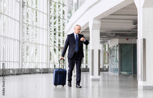 business trip, corporate and people concept - senior businessman walking with travel bag along office building or airport and checking time
