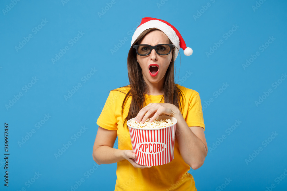 Shocked woman Santa girl in Christmas hat 3d imax glasses posing isolated on blue background. New Year 2020 celebration holiday concept. Mock up copy space. Watching movie film hold bucket of popcorn.
