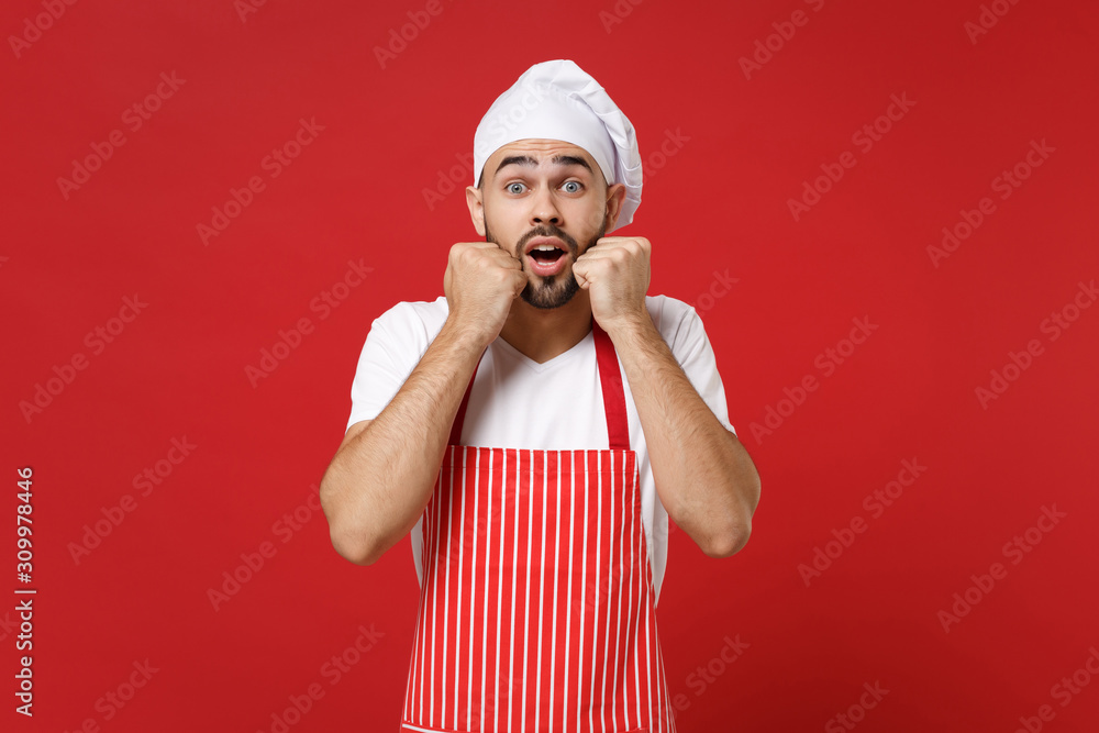 Shocked young bearded male chef cook or baker man in striped apron white t-shirt toque chefs hat posing isolated on bright red background. Cooking food concept. Mock up copy space. Clenching fists. Shocked young bearded male chef cook or baker man in striped apron white t-shirt toque chefs hat posing isolated on bright red background. Cooking food concept. Mock up copy space. Clenching fists.