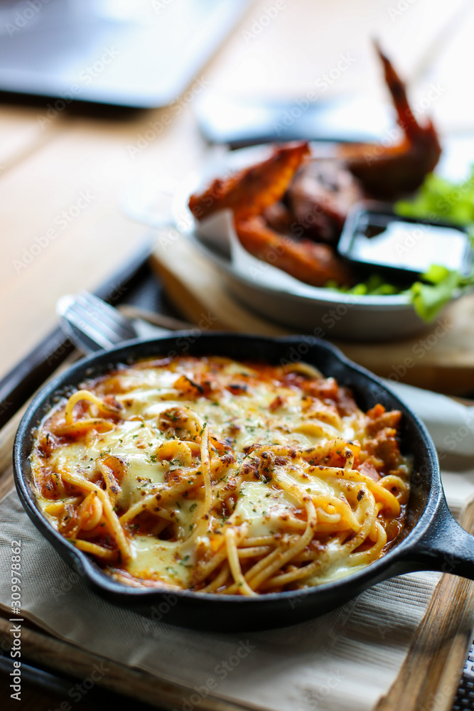 Cheesy spaghetti bolognese serve on pan metal top wood tray with fork and spoon side have chicken wing bbq background Cheesy spaghetti bolognese serve on pan metal top wood tray with fork and spoon side have chicken wing bbq background