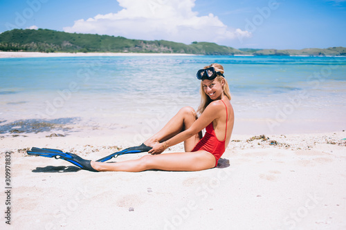 Cheerful adult female swimmer in bikini getting ready with snorkeling equipment on beach