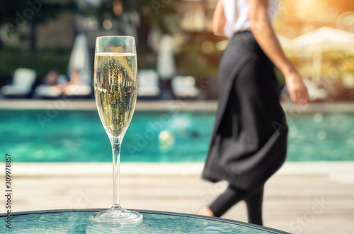 Close-up white champagne or prosecco glass against poolside at luxury resort hotel during vacation. Sparkling wine with rising bubbles with blue pool background outdoor. Waiter in black skirt leaving photo