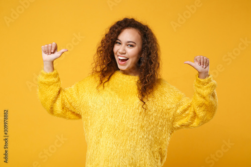 Cheerful funny young african american girl in fur sweater posing isolated on yellow orange wall background, studio portrait. People lifestyle concept. Mock up copy space. Pointing thumbs on herself.