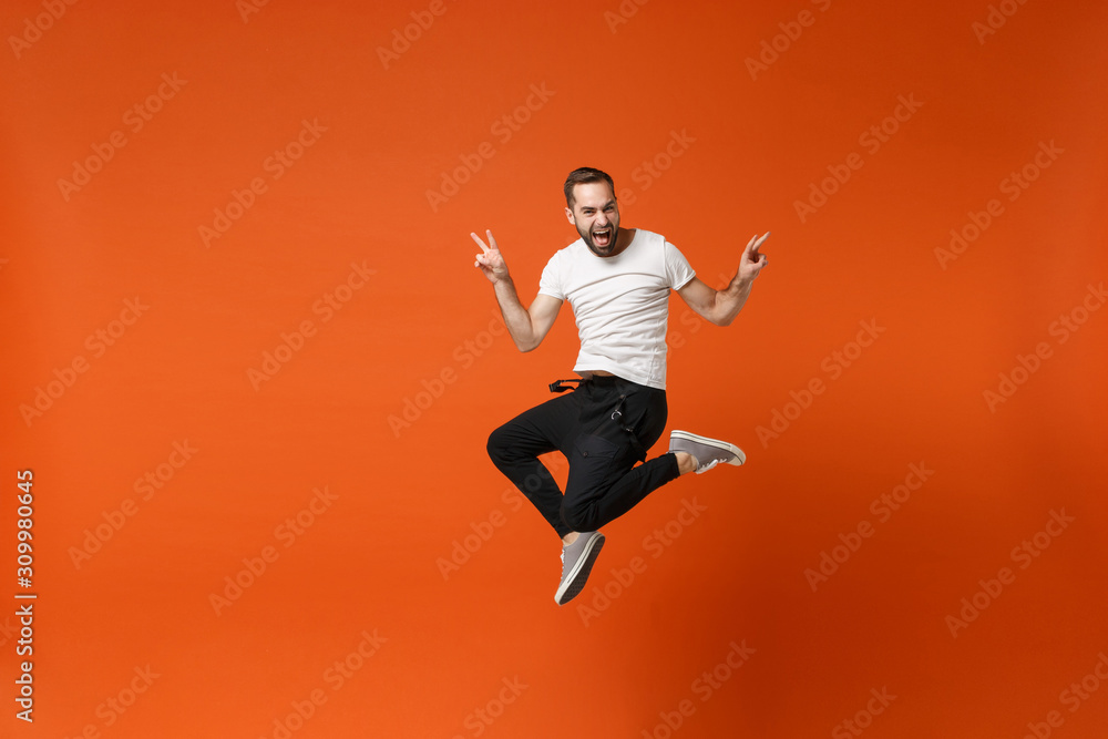 Funny young man in casual white t-shirt posing isolated on orange background studio portrait. People sincere emotions lifestyle concept. Mock up copy space. Having fun jumping, showing victory sign.