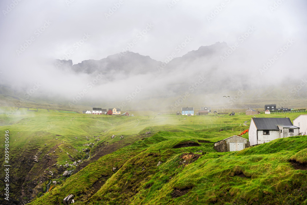 Fototapeta premium Foggy morning in Vidareidi village with white wooden houses surrounded with green hills. Faroe Islands, Denmark.
