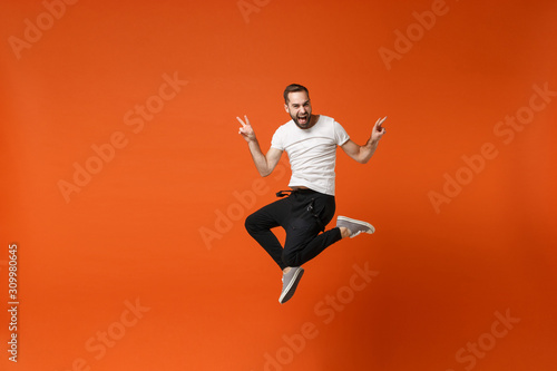 Funny young man in casual white t-shirt posing isolated on orange background studio portrait. People sincere emotions lifestyle concept. Mock up copy space. Having fun jumping, showing victory sign.