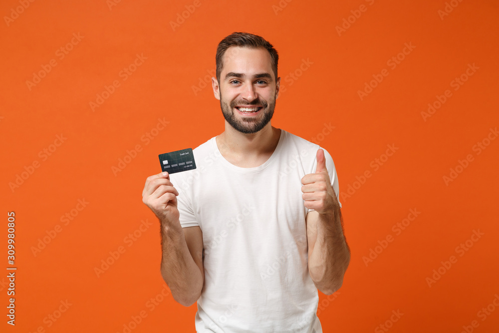 Smiling young man in casual white t-shirt posing isolated on bright orange wall background, studio portrait. People lifestyle concept. Mock up copy space. Holding credit bank card, showing thumb up. Smiling young man in casual white t-shirt posing isolated on bright orange wall background, studio portrait. People lifestyle concept. Mock up copy space. Holding credit bank card, showing thumb up.