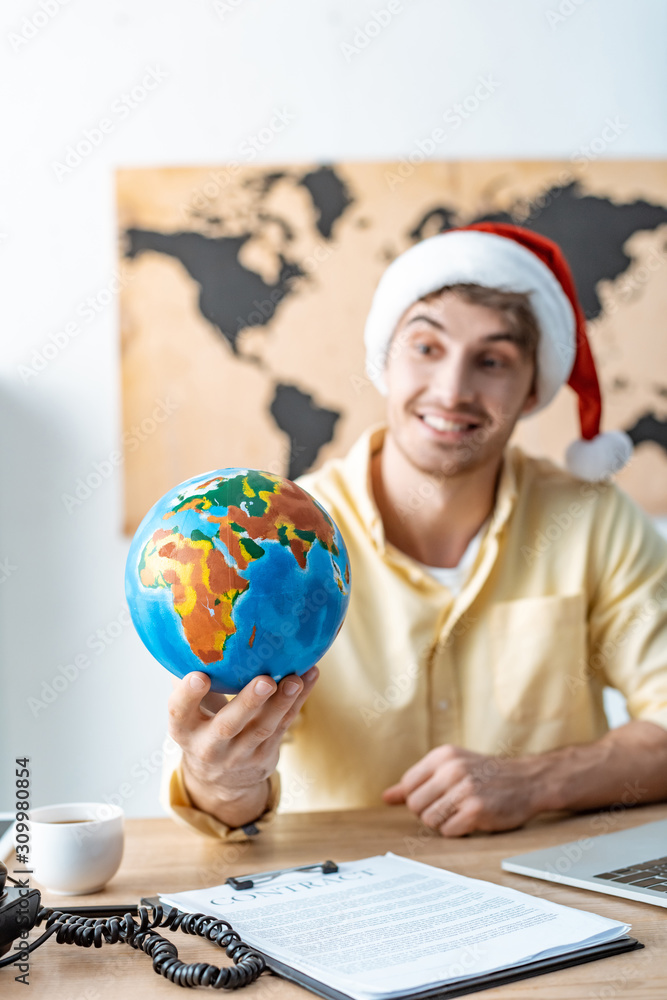 selective focus of smiling travel agent in santa hat holding globe while sitting at workplace selective focus of smiling travel agent in santa hat holding globe while sitting at workplace