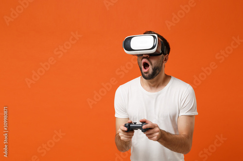 Shocked young man in casual white t-shirt posing isolated on orange background in studio. People lifestyle concept. Mock up copy space. Watching in headset of virtual reality, play game with joystick.
