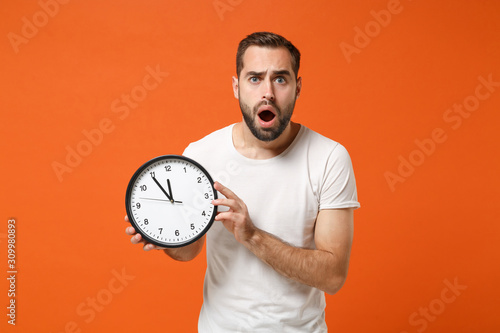 Shocked young man in casual white t-shirt posing isolated on bright orange wall background studio portrait. People sincere emotions lifestyle concept. Mock up copy space. Holding in hand round clock.