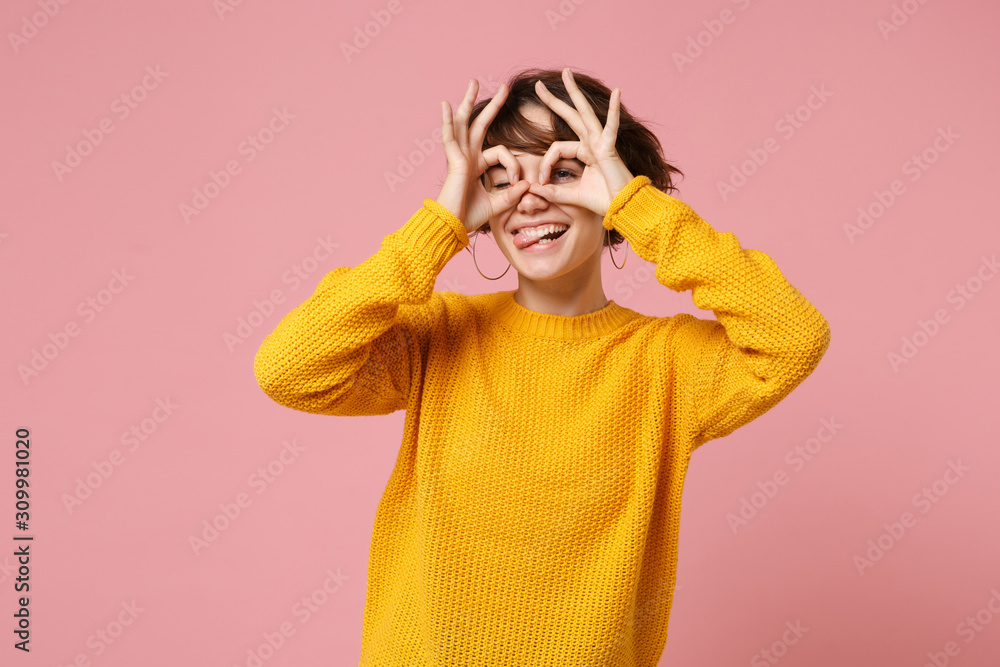 Funny young woman girl in yellow sweater posing isolated on pink wall background. People lifestyle concept. Mock up copy space. Holding hands near eyes imitating binoculars or glasses showing tongue. Funny young woman girl in yellow sweater posing isolated on pink wall background. People lifestyle concept. Mock up copy space. Holding hands near eyes imitating binoculars or glasses showing tongue.