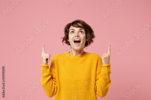 Amazed young brunette woman girl in yellow sweater posing isolated on pastel pink background, studio portrait. People sincere emotions lifestyle concept. Mock up copy space. Pointing index fingers up.