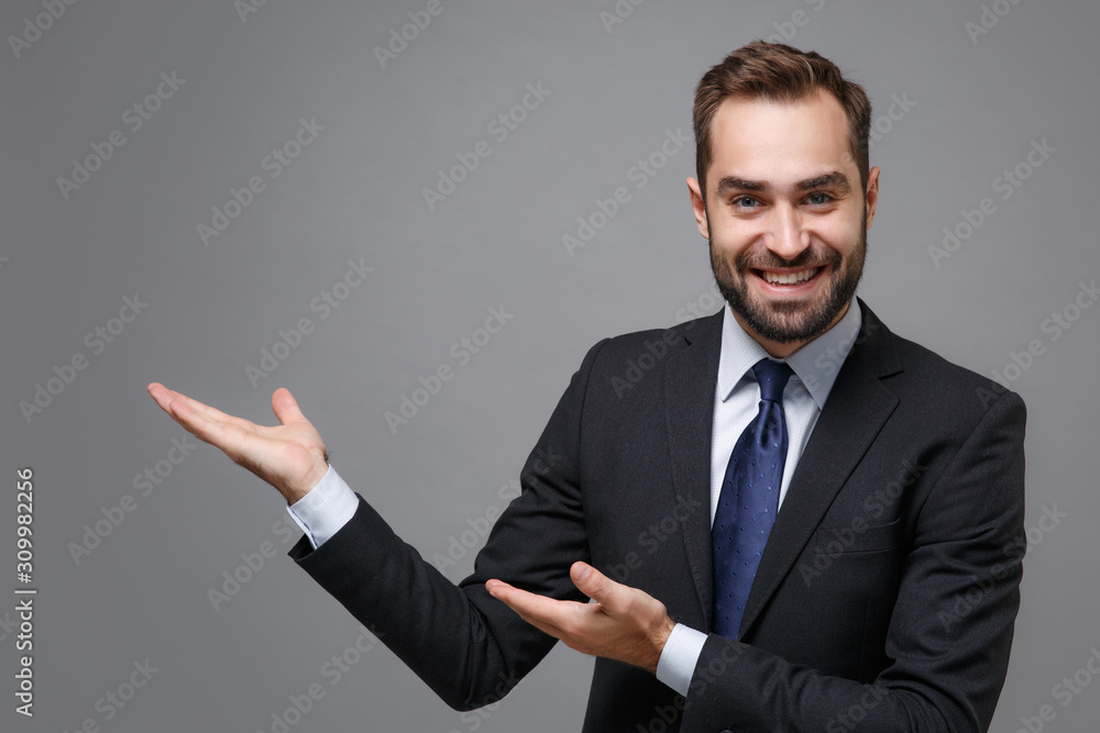 Smiling young bearded business man in classic black suit shirt tie posing isolated on grey background in studio. Achievement career wealth business concept. Mock up copy space. Pointing hands aside. Smiling young bearded business man in classic black suit shirt tie posing isolated on grey background in studio. Achievement career wealth business concept. Mock up copy space. Pointing hands aside.