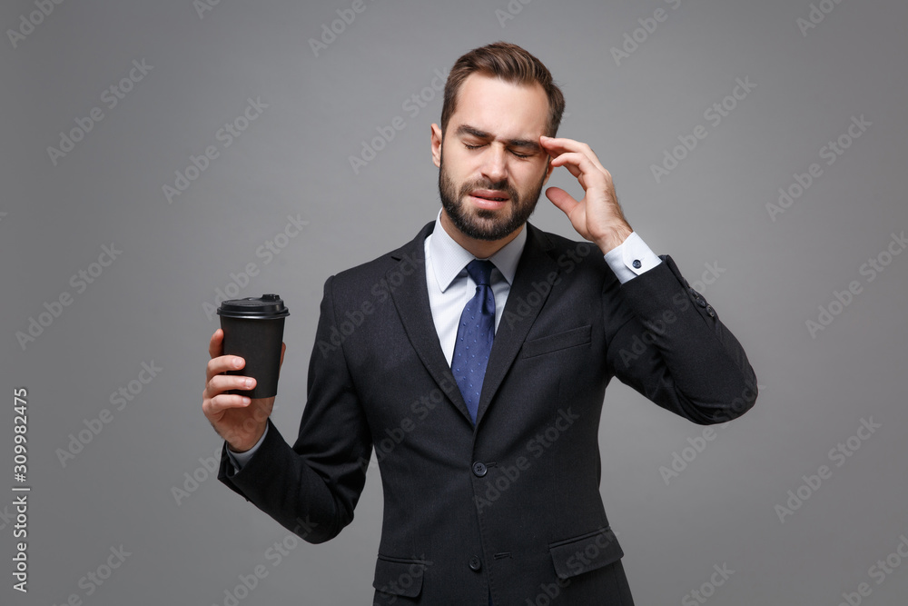 Sick young business man in classic black suit shirt tie posing isolated on grey background. Achievement career wealth business concept. Mock up copy space. Hold cup of coffee or tea put hand on head. Sick young business man in classic black suit shirt tie posing isolated on grey background. Achievement career wealth business concept. Mock up copy space. Hold cup of coffee or tea put hand on head.