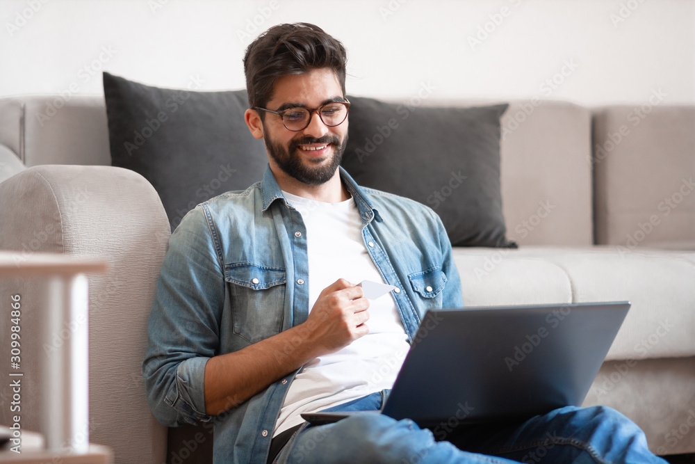 Smiling caucasian hipster sitting on floor in living room, holding laptop in lap and using credit card for online shopping or paying bills. Smiling caucasian hipster sitting on floor in living room, holding laptop in lap and using credit card for online shopping or paying bills.