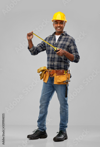 profession, construction and building - happy smiling indian worker or builder in helmet with ruler and tool belt over grey background