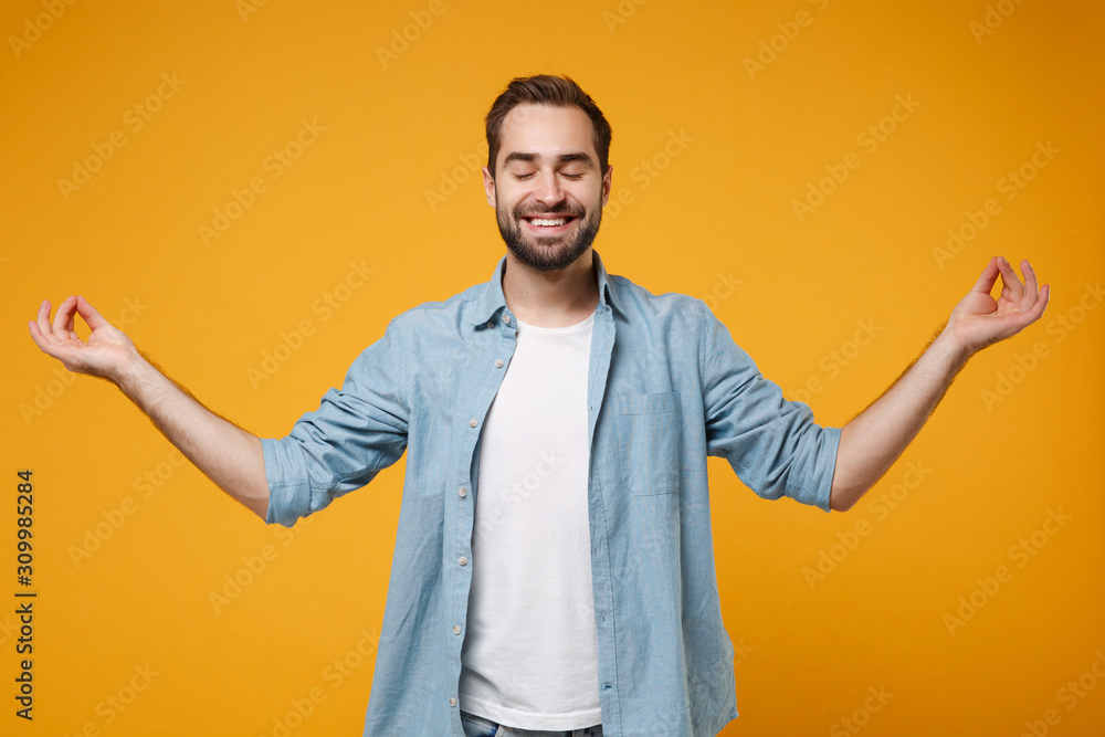 Smiling young bearded man in blue shirt posing isolated on yellow orange background. People lifestyle concept. Mock up copy space. Hold hands in yoga gesture, relaxing meditating, keeping eyes closed. Smiling young bearded man in blue shirt posing isolated on yellow orange background. People lifestyle concept. Mock up copy space. Hold hands in yoga gesture, relaxing meditating, keeping eyes closed.