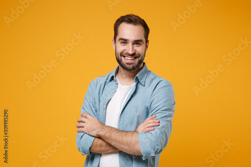 Smiling young bearded man in casual blue shirt posing isolated on yellow orange wall background, studio portrait. People sincere emotions lifestyle concept. Mock up copy space. Holding hands crossed.