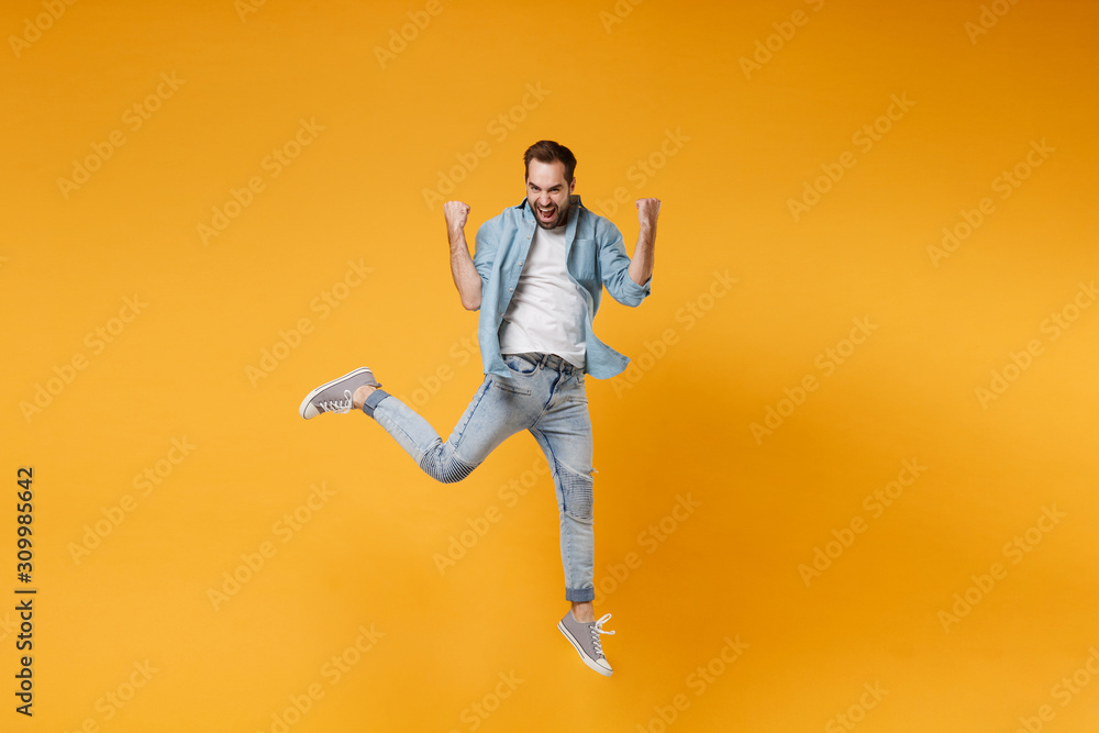 Joyful young bearded man in casual blue shirt posing isolated on yellow orange background, studio portrait. People sincere emotions lifestyle concept. Mock up copy space. Jumping doing winner gesture.