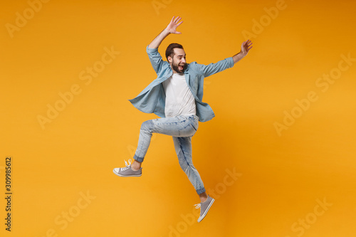 Joyful young bearded man in casual blue shirt posing isolated on yellow orange wall background studio portrait. People sincere emotions lifestyle concept. Mock up copy space. Jumping, rising hands up.