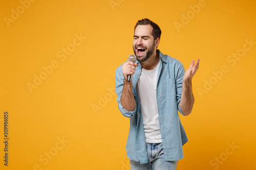 Handsome young man in casual blue shirt posing isolated on yellow orange wall background, studio portrait. People lifestyle concept. Mock up copy space. Sing song in microphone, keeping eyes closed.